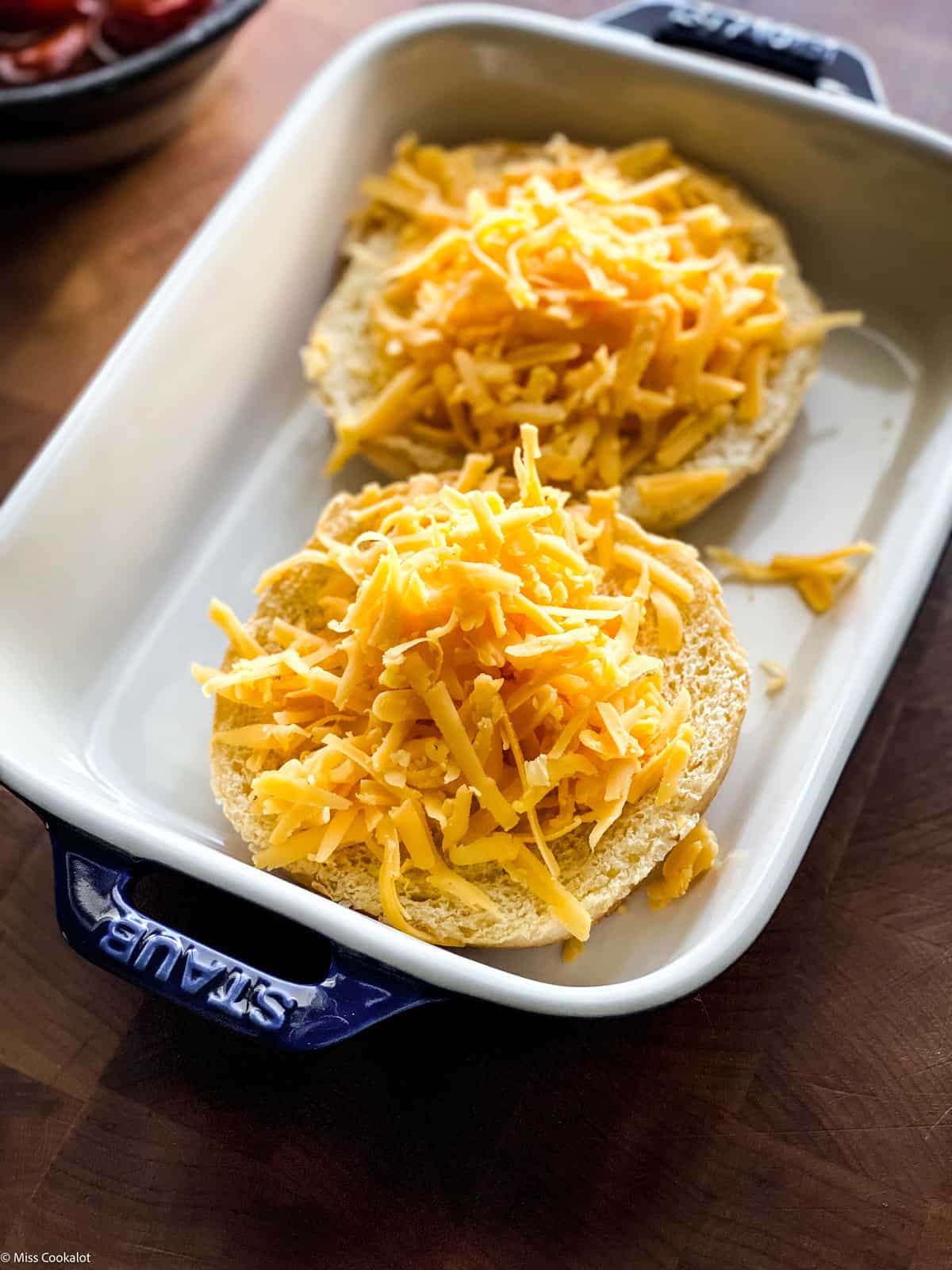 Two brioche bun halves topped with shredded cheddar cheese in a baking pan.