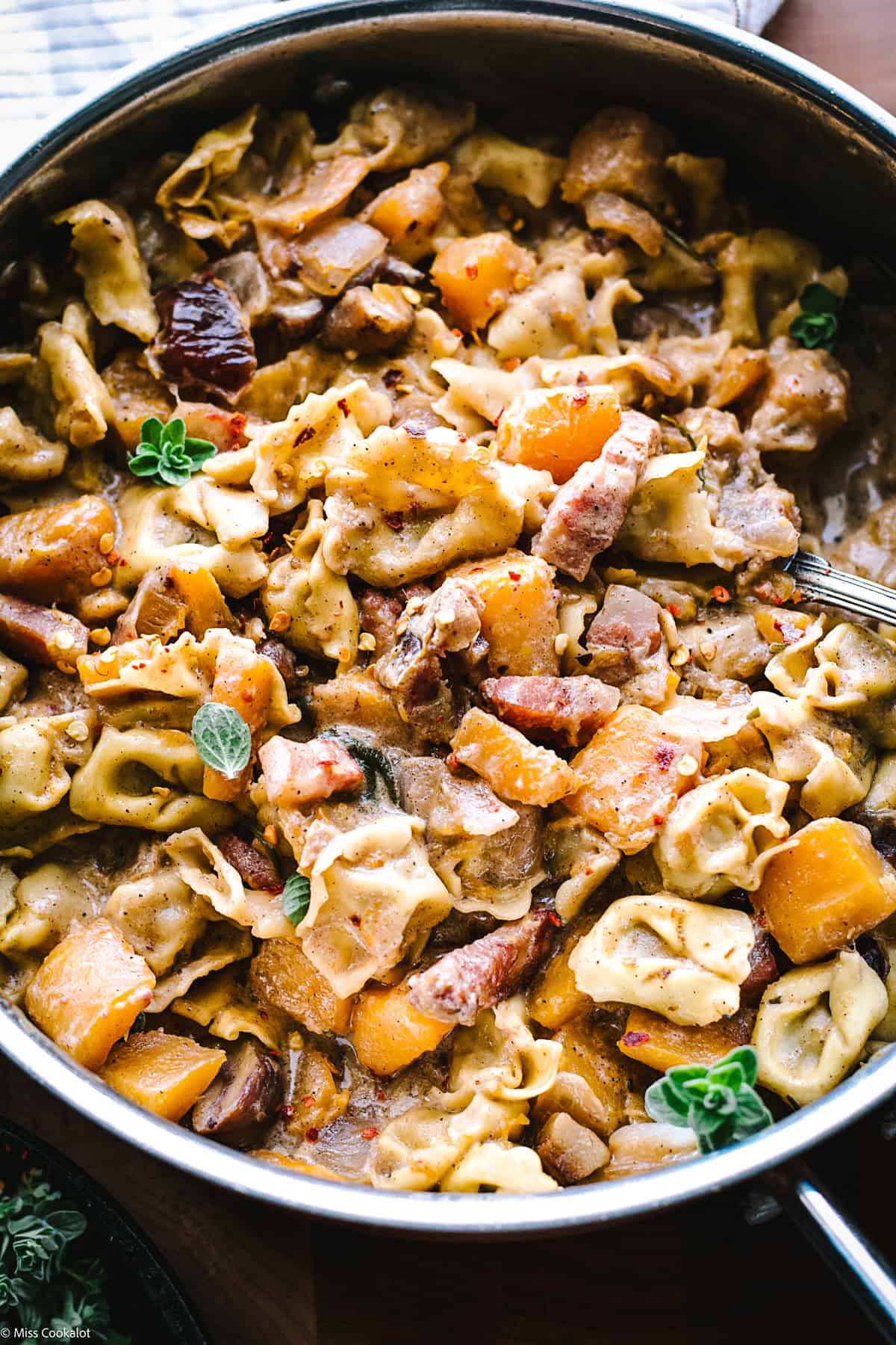 A skillet with pumpkin stew with tortellini and a spoon, with herbs and spices on a table, overview.