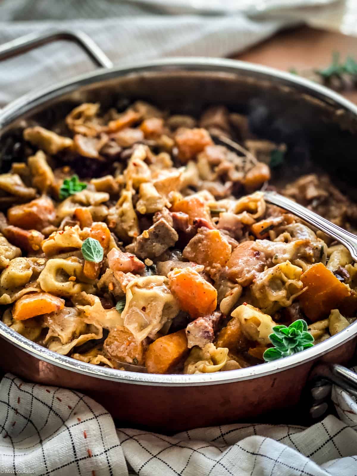 A deep large sauce pan pumpkin stew with tortellini and a spoon, a napkin and a plate with herbs and spices.