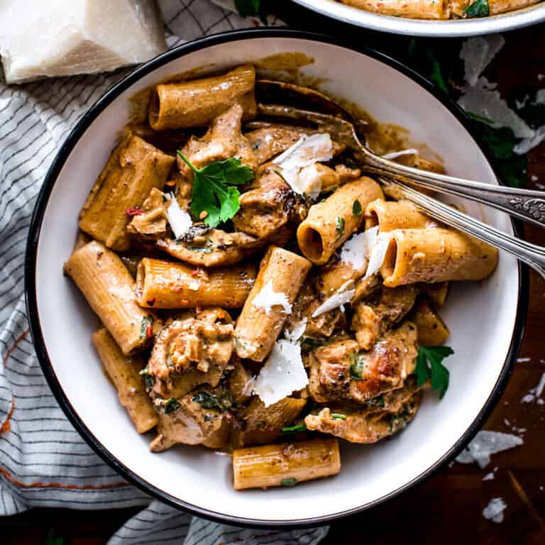 Overview of a plate with chipotle chicken pasta, a fork and a spoon, a cloth napkin and a pice of parmesan with some fresh parsley and partial view of another plate.