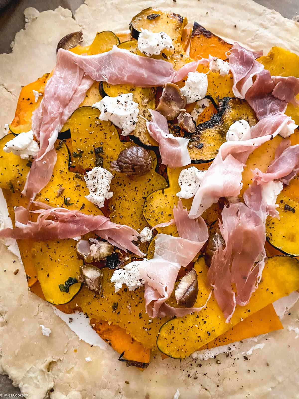 A galette crust with goat cheese squash, prosciutto and chestnuts on parchment paper.