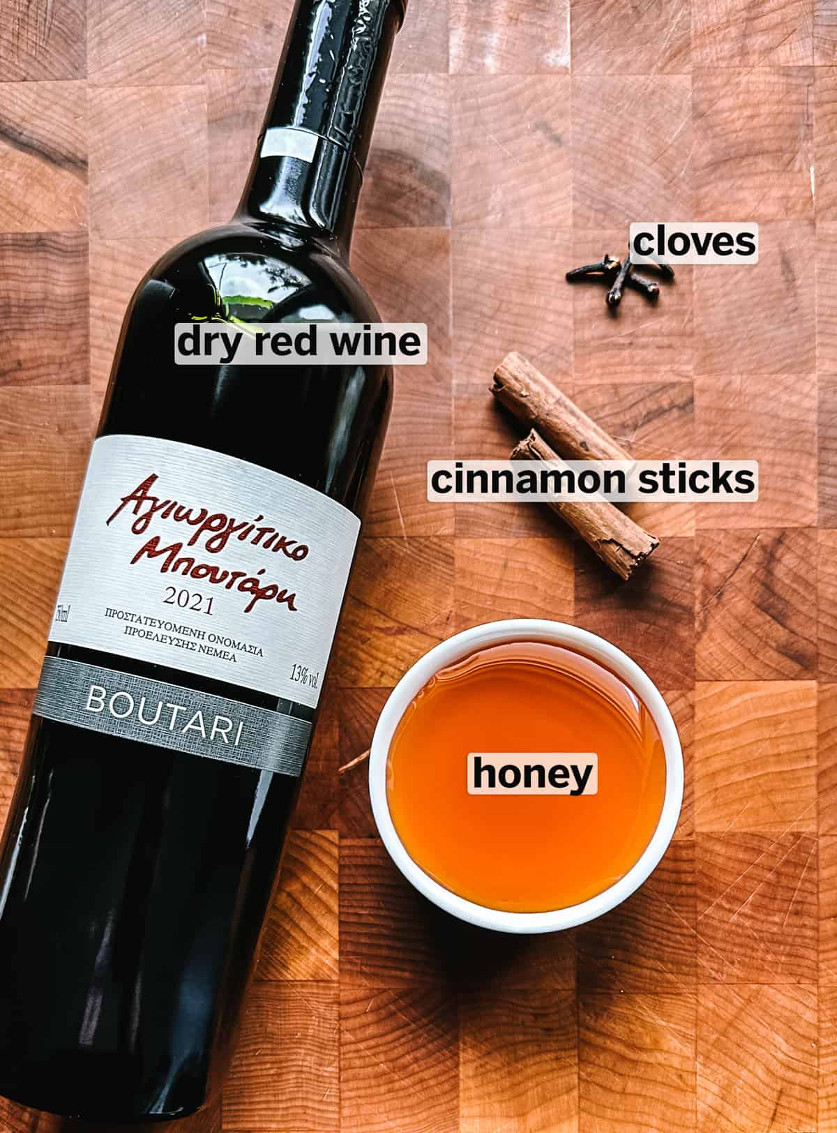 A bottle of red wine, a bowl with honey , cinnamon sticks and cloves on a wooden surface.